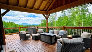 Une terrasse couverte, aménagée avec des meubles en osier, une table et offrant une vue sur une forêt et des montagnes.