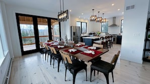 Un espace repas moderne avec une longue table dressée pour six personnes, des chaises en bois et une vue sur l’extérieur grâce à de grandes fenêtres.