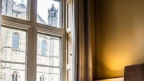 Habitación de hotel con cama, lámpara y vista a un edificio histórico a través de amplios ventanales.