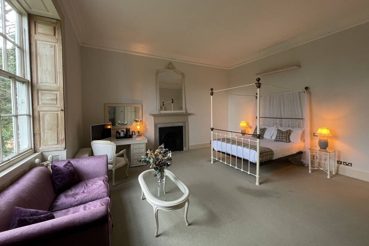 A bedroom with a four-poster bed, a sitting area with a purple sofa, a desk with a mirror, and a fireplace.