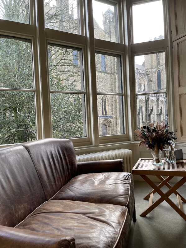 Une pièce avec un canapé en cuir marron, un radiateur et une table sur laquelle est posé un vase de fleurs. De grandes fenêtres offrent une vue sur un bâtiment historique.