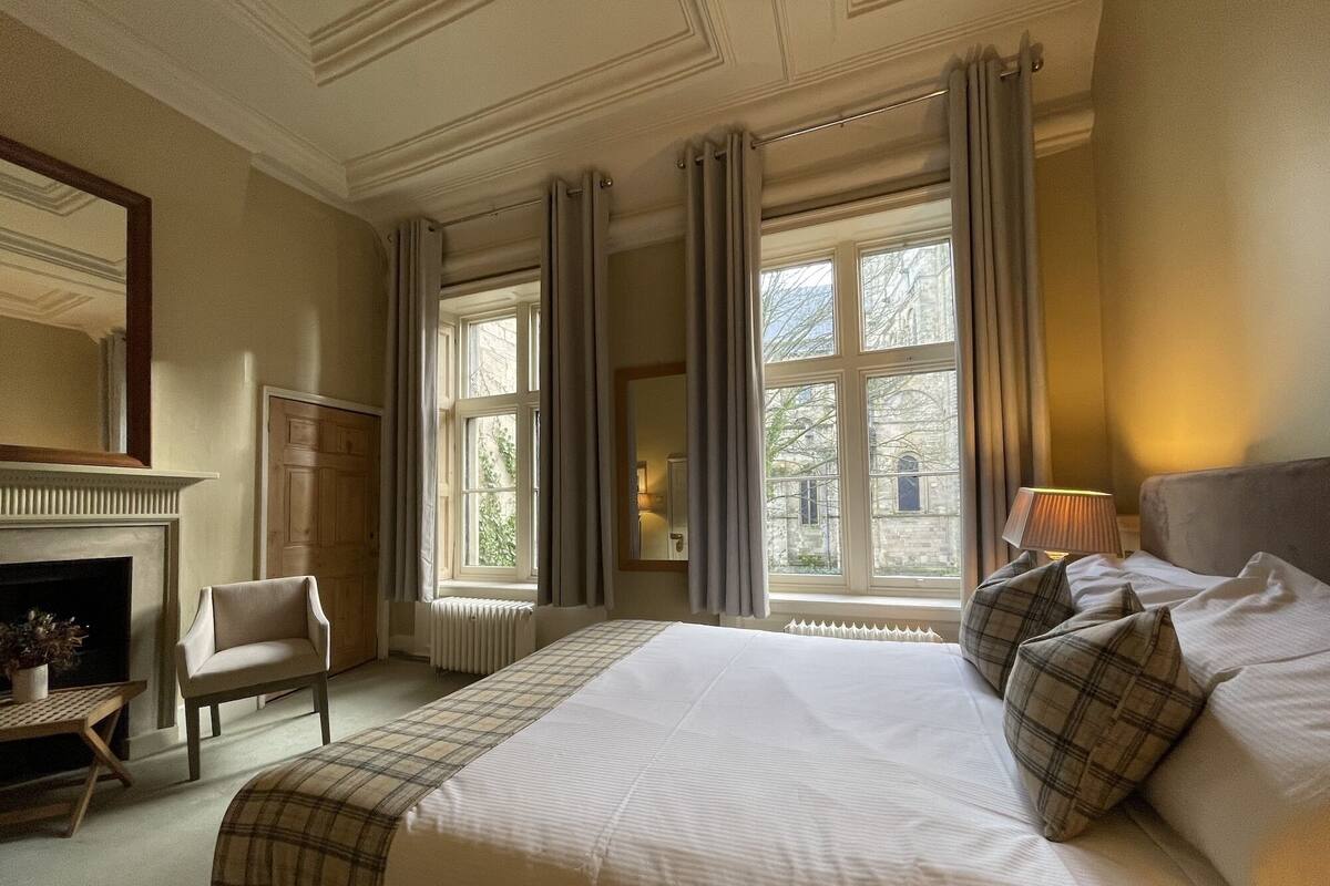 A hotel room with a large bed, a chair, a fireplace, and large windows with curtains.