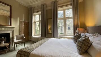 A hotel room with a large bed, a chair, a fireplace, and large windows with curtains.