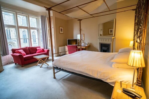 A four-poster bed with a white mattress, a red sofa, a small table, and a lamp on a bedside table. - The Old Deanery (Ripon)