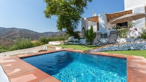 A swimming pool with a clear blue water, surrounded by a stone border. There are two lounge chairs and a tree nearby. The background features a white building with a red roof and a view of the hills. - Cortijo Horizonte Solset (Frigiliana)