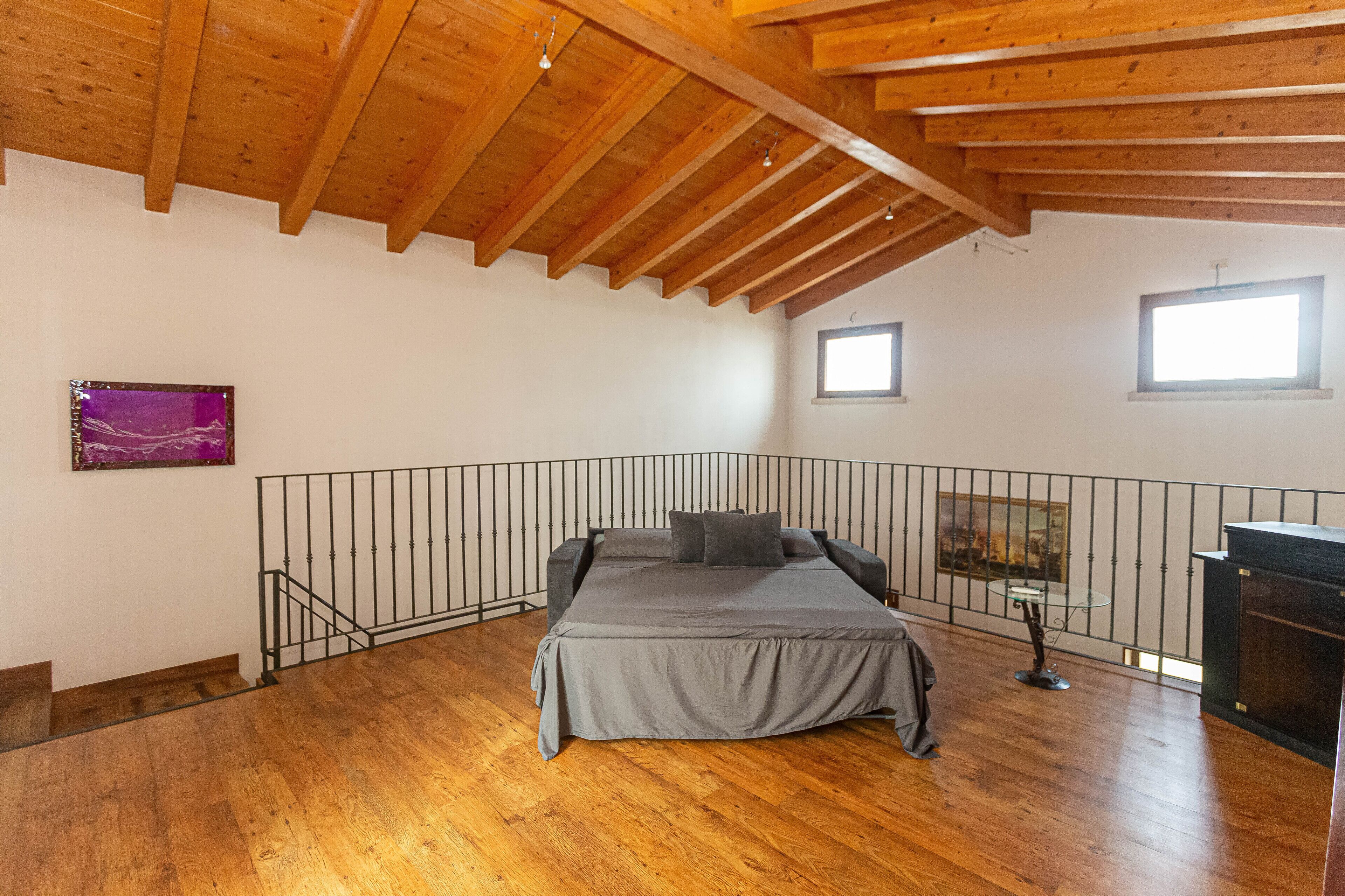 Une chambre avec un plafond en bois, un lit avec une literie grise, deux fenêtres et un tableau au mur.