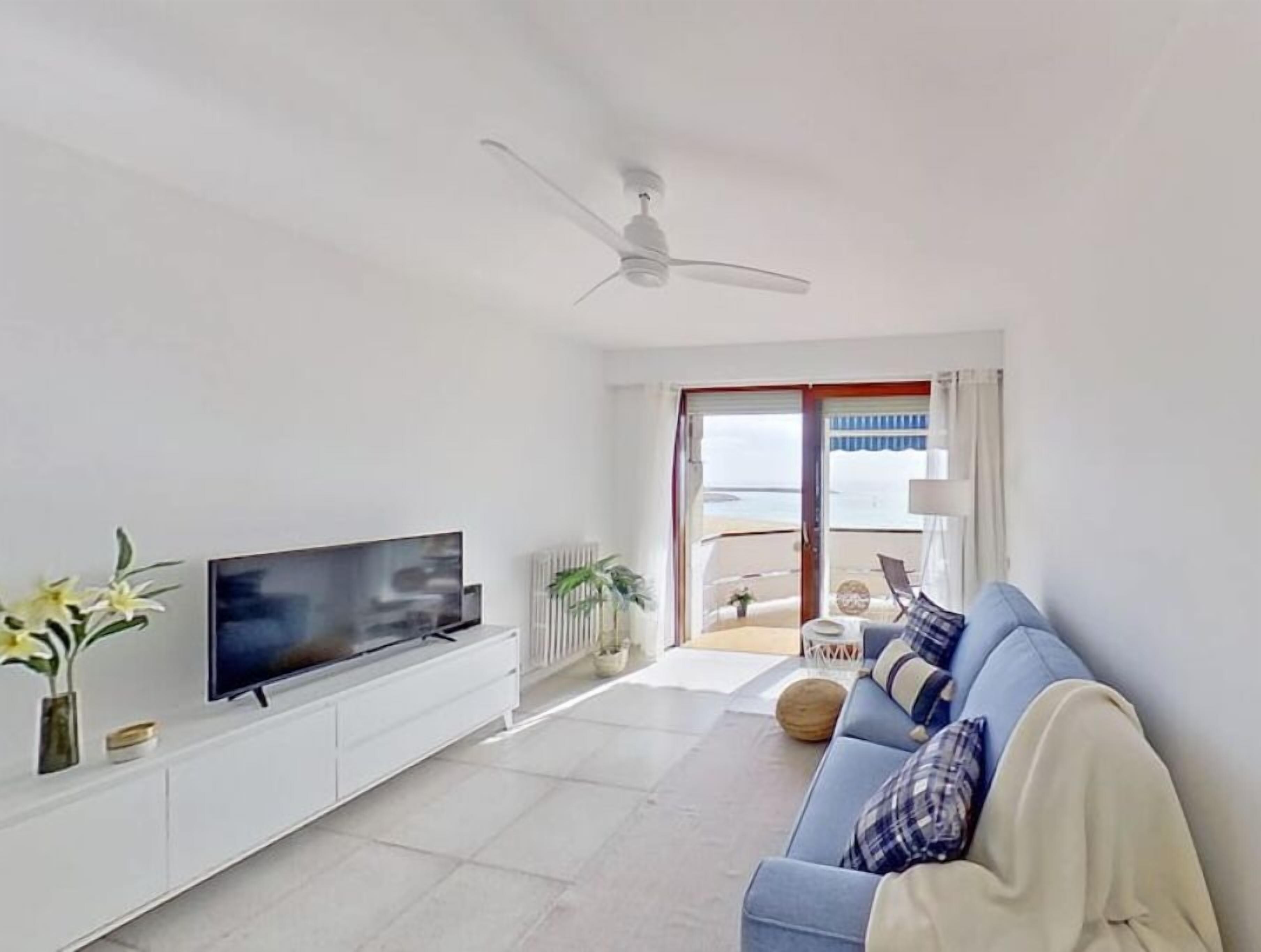 Una sala de estar moderna con televisor de pantalla plana, ventilador de techo, sofá y vista a la playa a través de la puerta corrediza de vidrio.