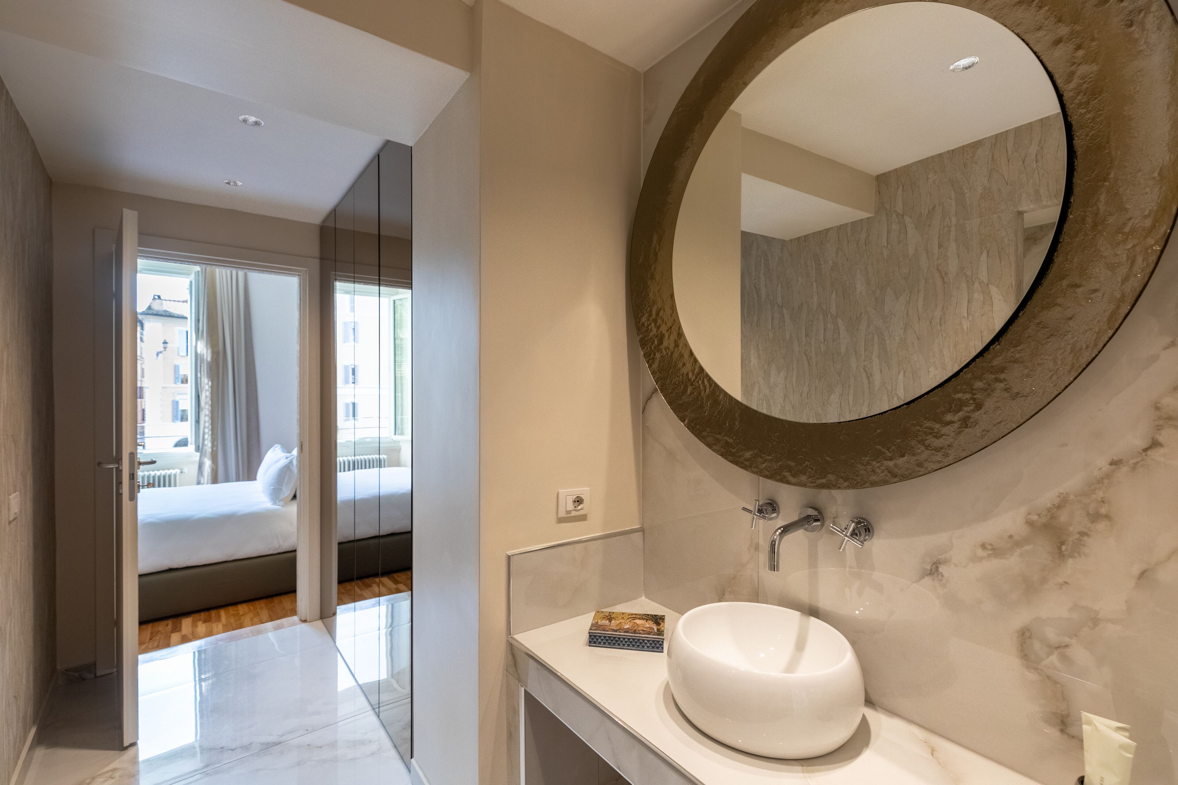 A modern bathroom with a large mirror, a round sink, and a view of a bedroom with a bed and a window.