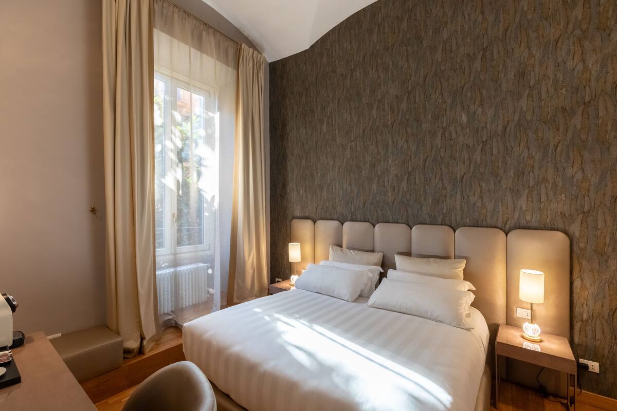 A hotel room with a large bed, two bedside tables with lamps, a desk with a chair, and a window with curtains.