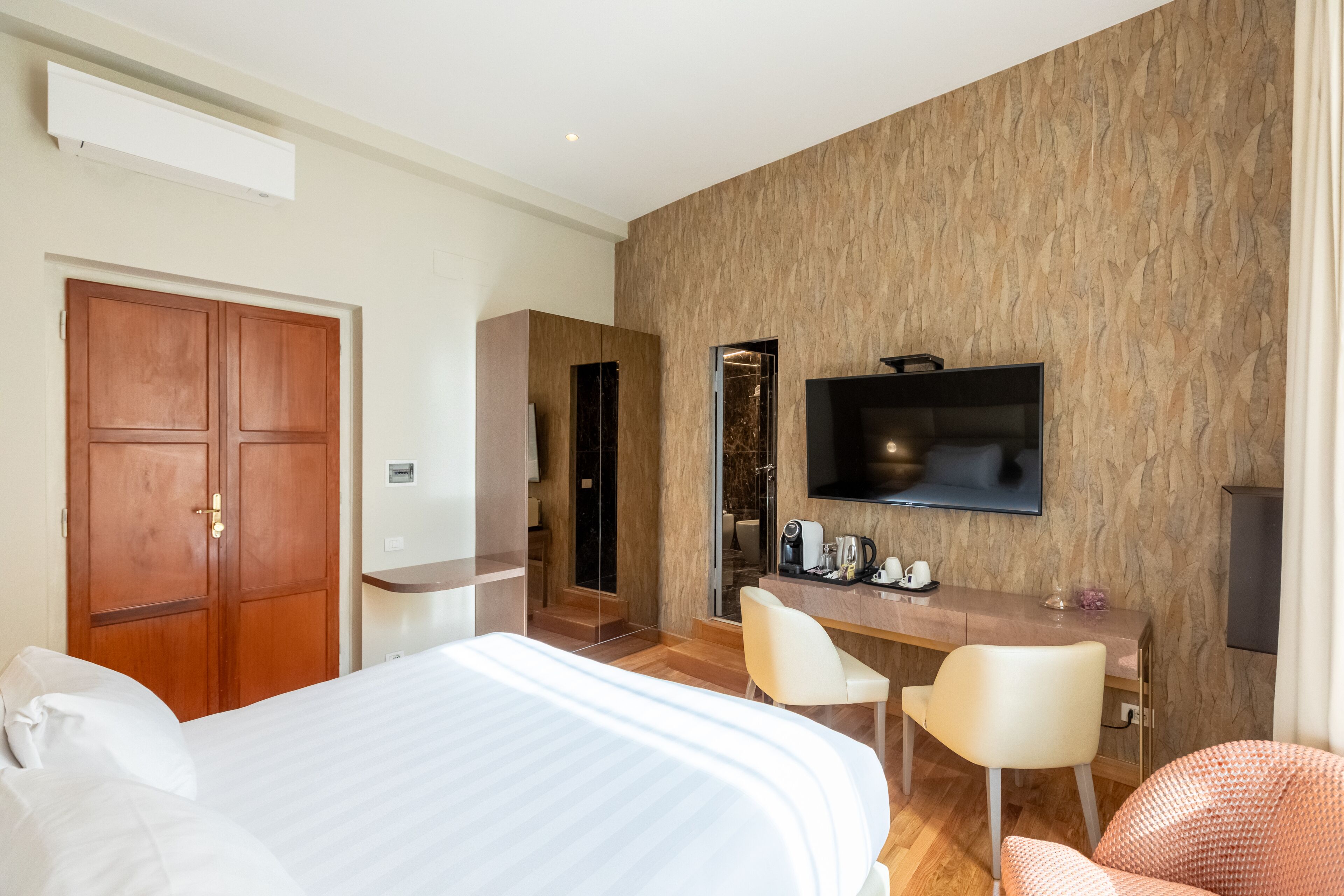 A modern hotel room with a large bed, a desk with two chairs, a flat-screen TV mounted on the wall, and a wooden wardrobe.