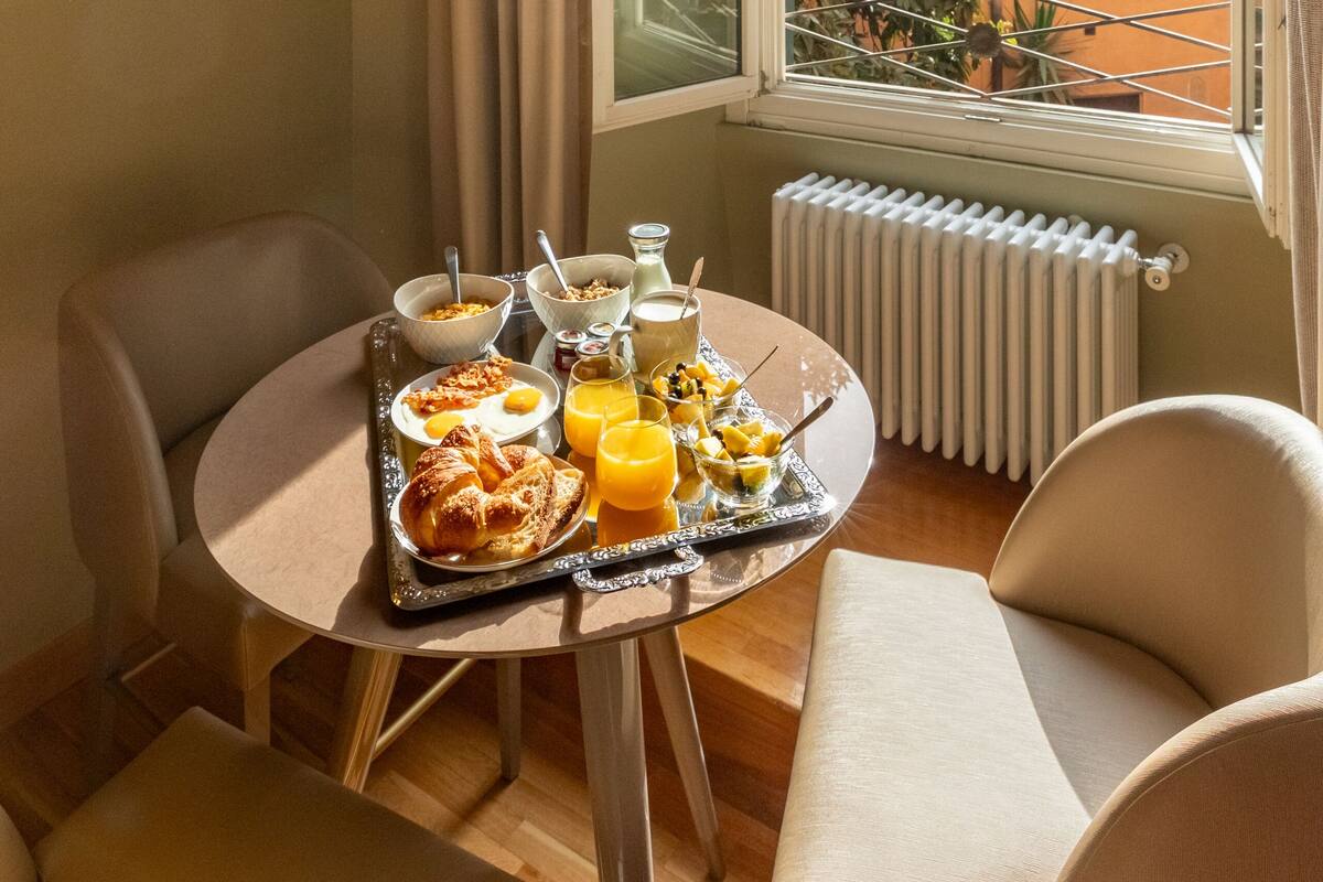 A breakfast table with croissants, orange juice, and cereal, set in a room with a view of buildings and greenery through a window.