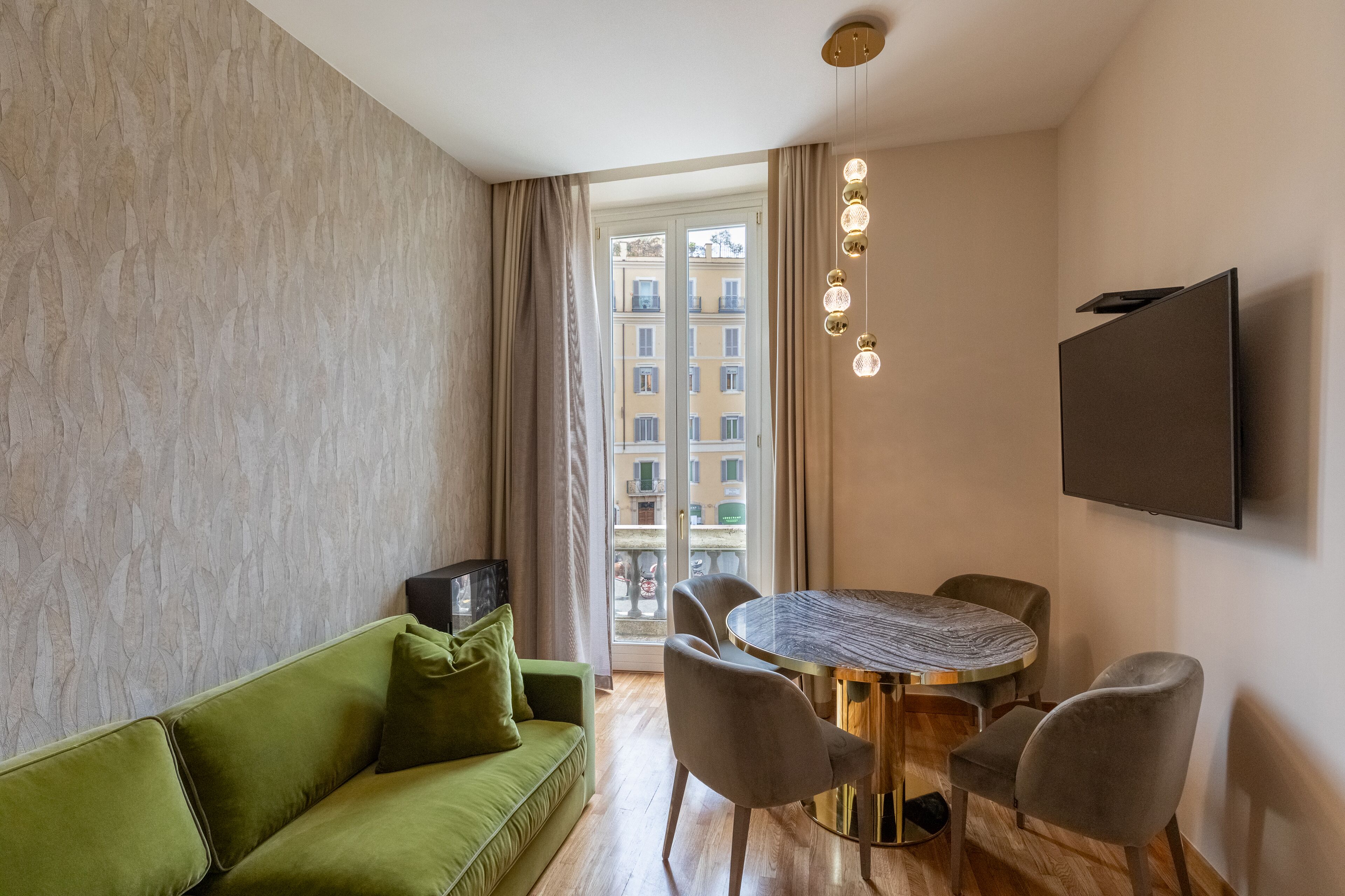 Una sala de estar moderna con un sofá verde, una mesa de comedor redonda con sillas, un televisor de pantalla plana montado en la pared y vistas a edificios a través de un ventanal.