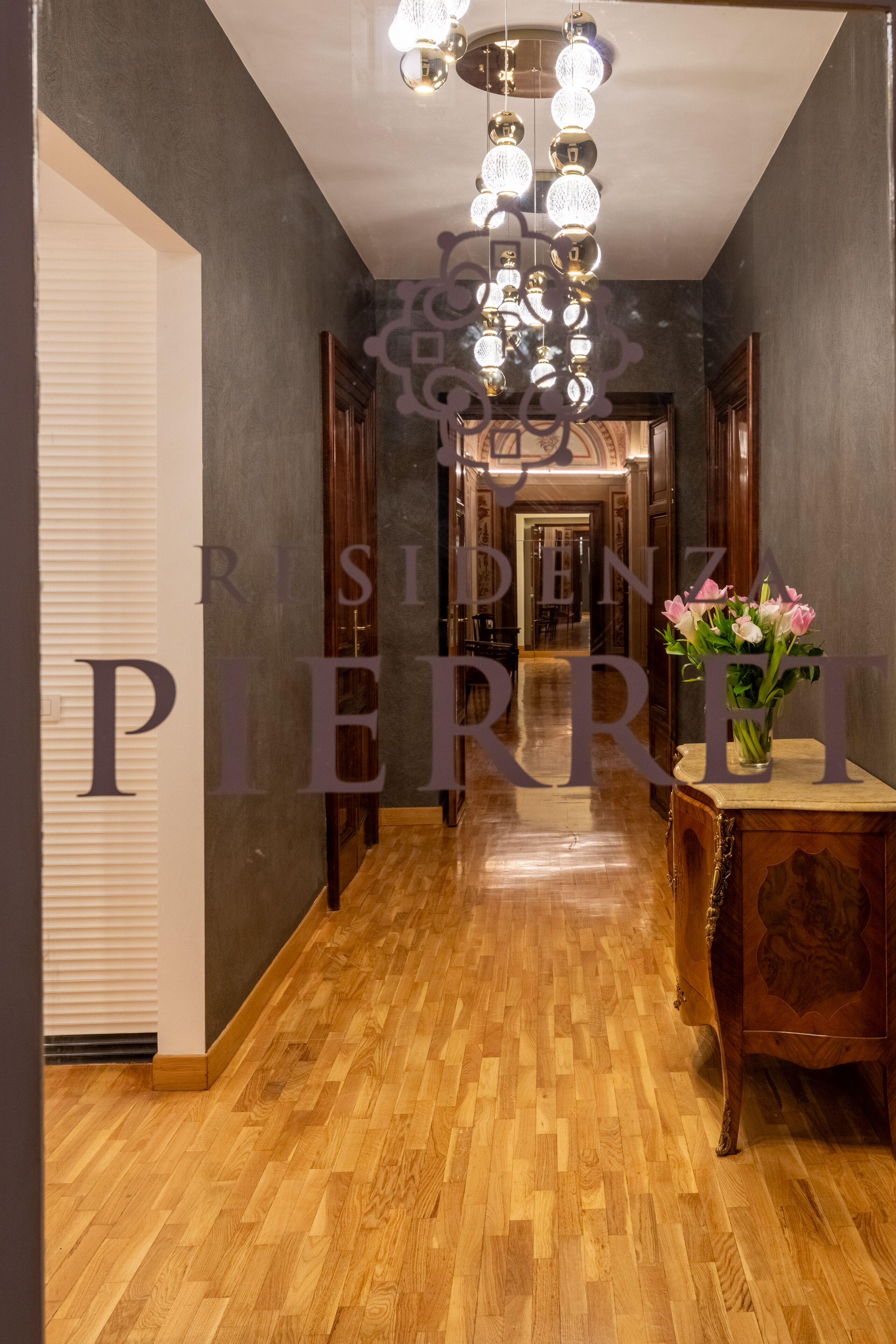 A hallway with a wooden floor, a wooden table with a vase of flowers, and a chandelier.