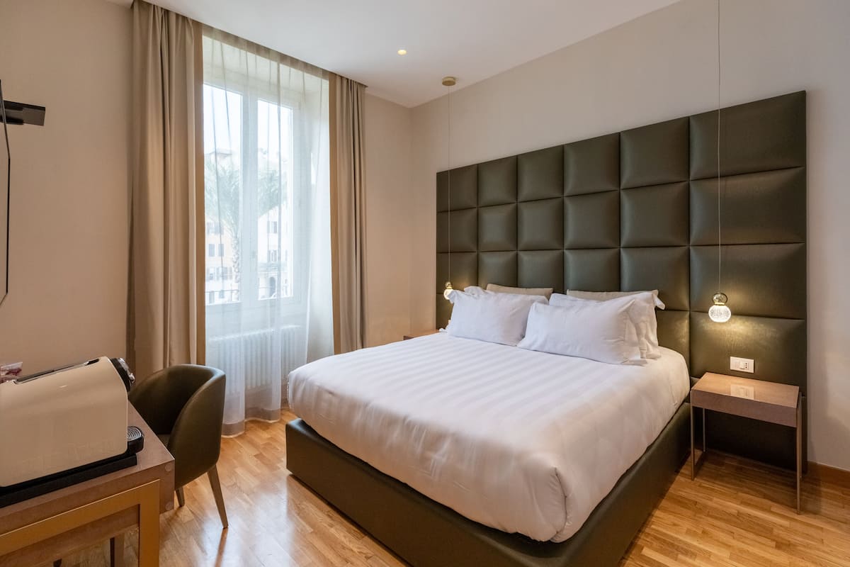 A hotel room with a large bed, a desk with a chair, a nightstand, and a window with curtains.