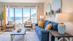 A room with a large window overlooking a beach, a blue sofa with yellow cushions, a wooden coffee table, and a lamp on a side table.