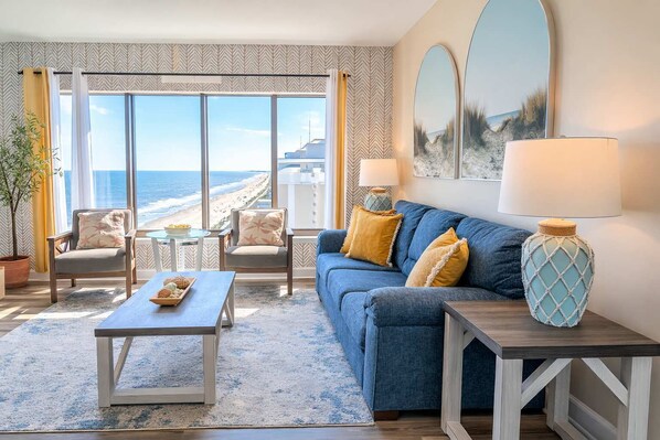 Una habitación con un ventanal grande con vistas a la playa, un sofá azul con cojines amarillos, una mesa de centro de madera y una lámpara en una mesita auxiliar.