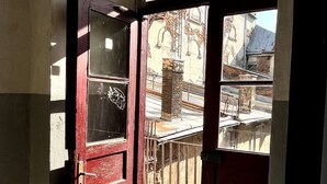 Uma porta vermelha aberta que dá para uma varanda com piso de azulejos e vista para edifícios e uma rua.