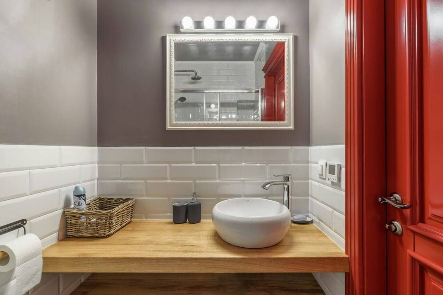 Un bagno con un lavandino bianco, uno specchio illuminato, una porta rossa e un piano d'appoggio in legno.
