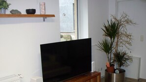 Una sala de estar con una televisión de pantalla plana sobre un mueble de madera, una planta en maceta y un estante con objetos decorativos.