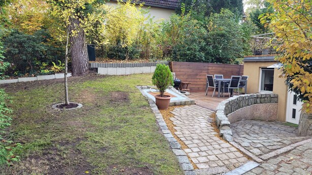 Un patio trasero con un adoquinado, una mesa pequeña y sillas, un árbol en maceta y un muro de piedra.
