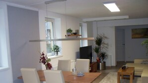 Una sala de estar moderna con comedor, que incluye una mesa de madera, sillas blancas, un televisor y plantas en macetas.