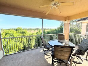 A covered balcony with a table and chairs, a ceiling fan, and a view of greenery.
