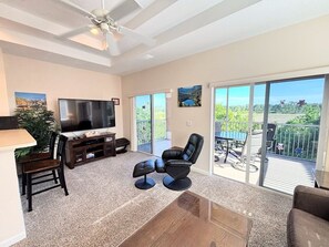 A living room with a TV, seating, a balcony with a table and chairs, and a view of greenery. - Right Direction Rentals 1 Bedroom Villa 8272 (Port St. Lucie)