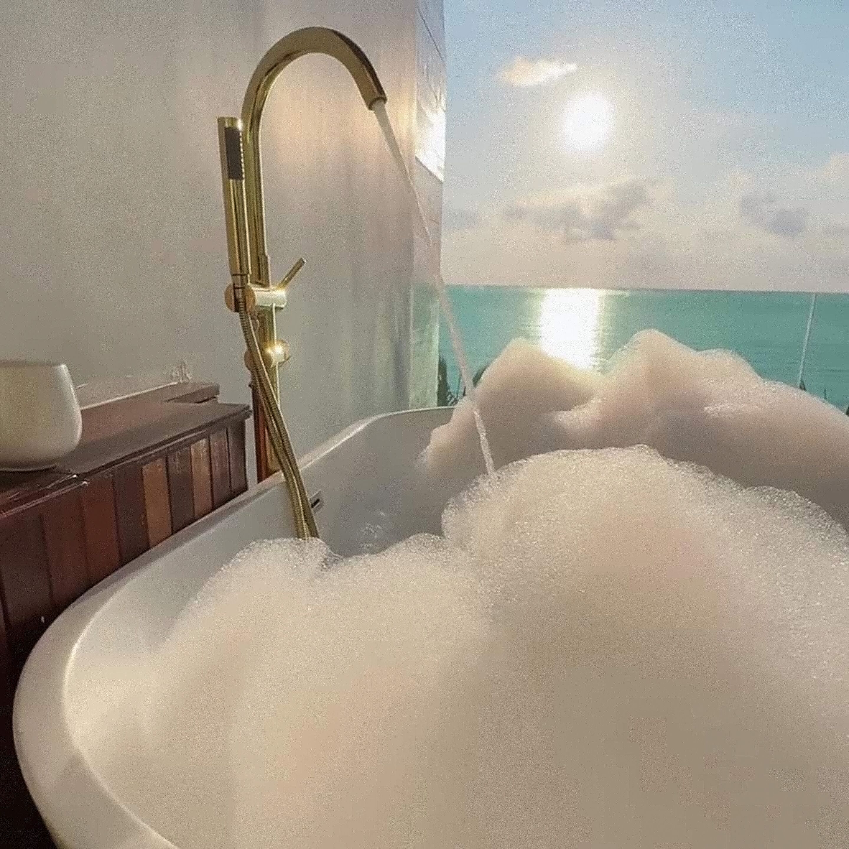 A bathtub filled with bubbles, a gold faucet, and a view of the ocean.