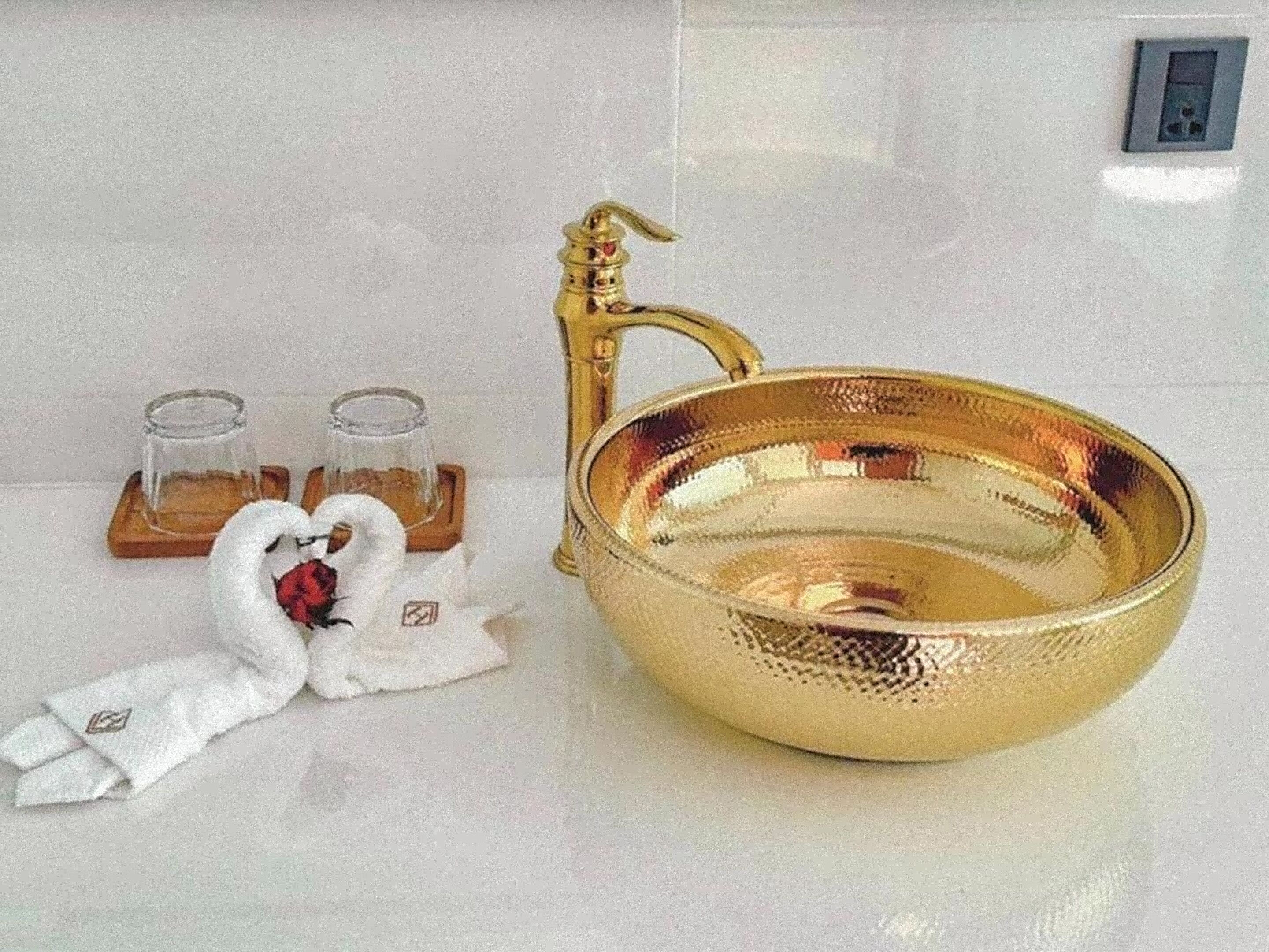 Une salle de bain avec un lavabo doré, deux verres en verre posés sur des sous-verres en bois, et une serviette blanche ornée d’une rose rouge.