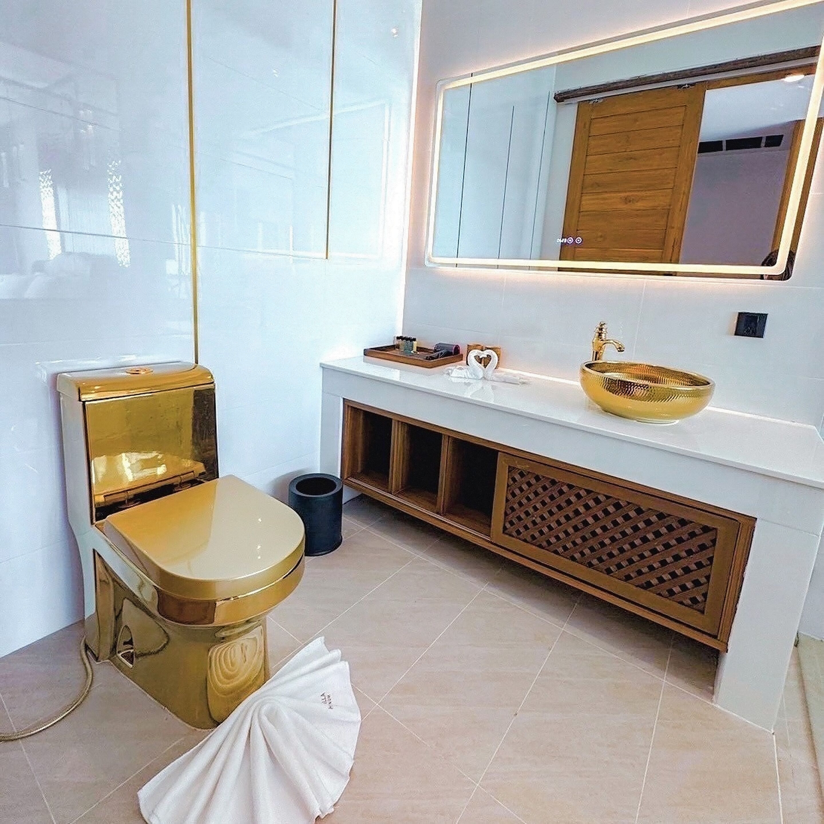 Un baño moderno con inodoro dorado, lavabo blanco con recipiente dorado y un espejo grande sobre el lavabo.
