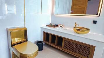 Un baño moderno con inodoro dorado, lavabo blanco con recipiente dorado y un espejo grande sobre el lavabo.