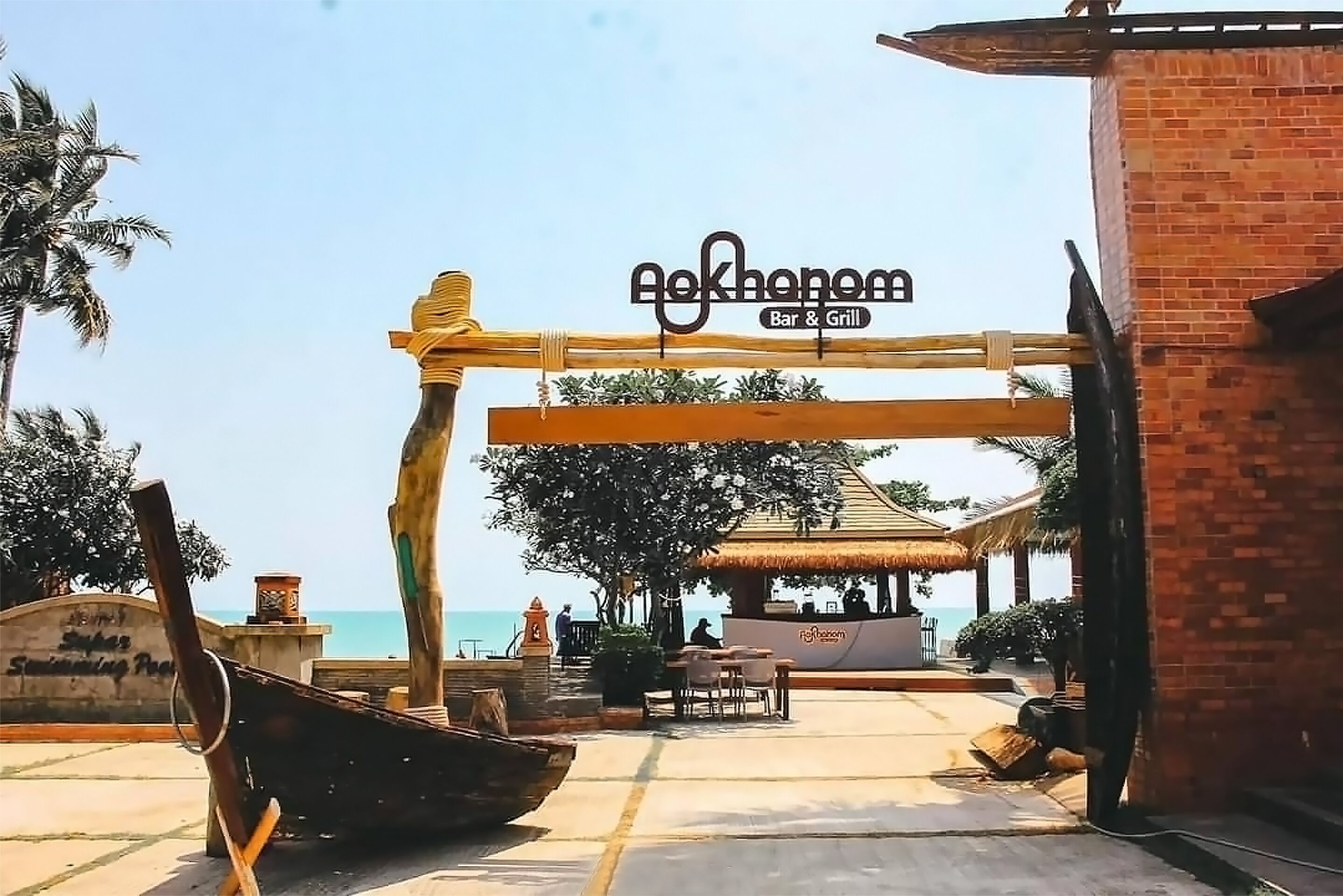 Una entrada frente a la playa a un bar y parrilla llamado 'Aekhonom', con una estructura de techo de paja, palmeras y vistas despejadas al mar.