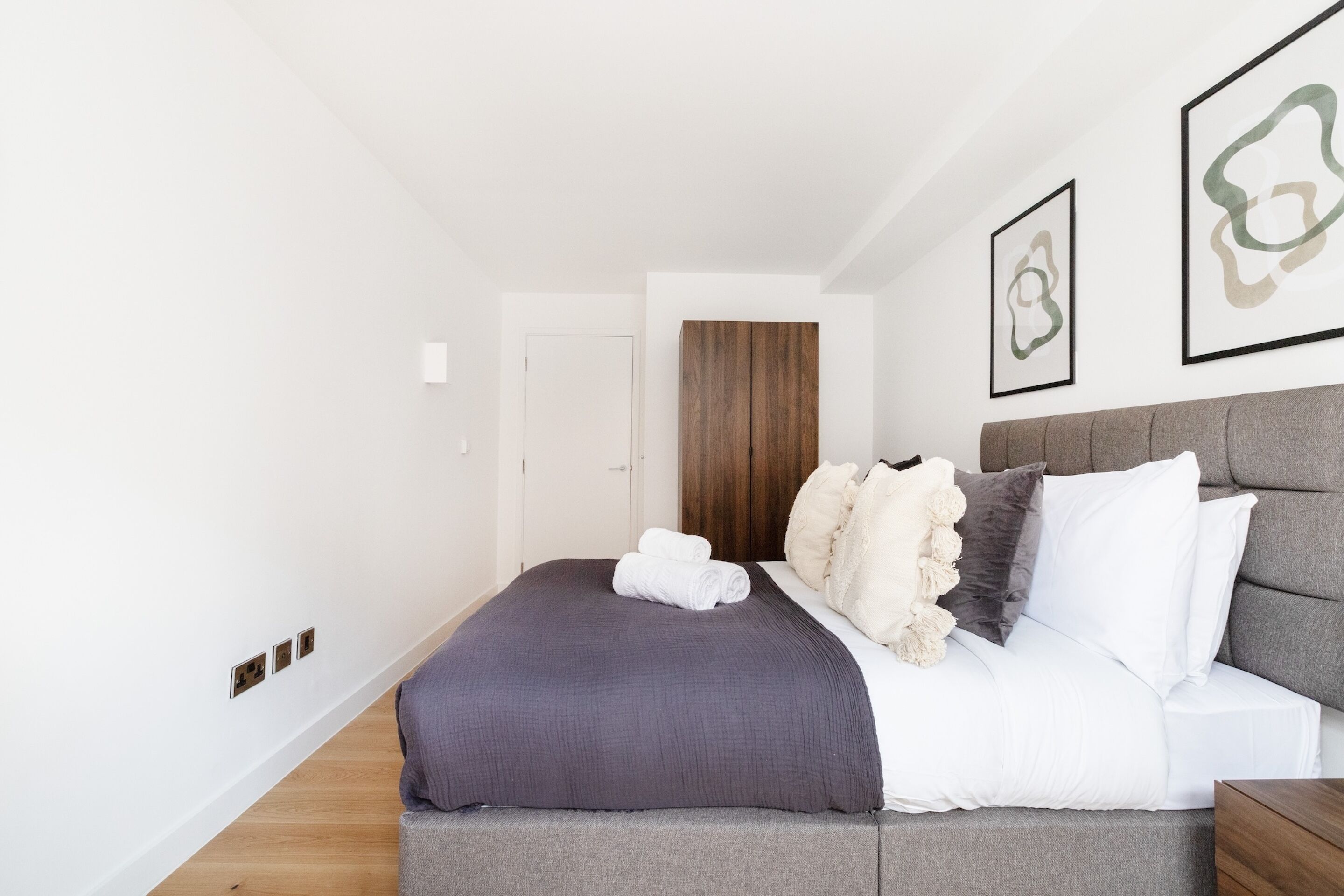 A modern bedroom with a grey bed, white pillows, and a grey blanket. There are framed abstract art pieces on the wall and a wooden wardrobe in the background.