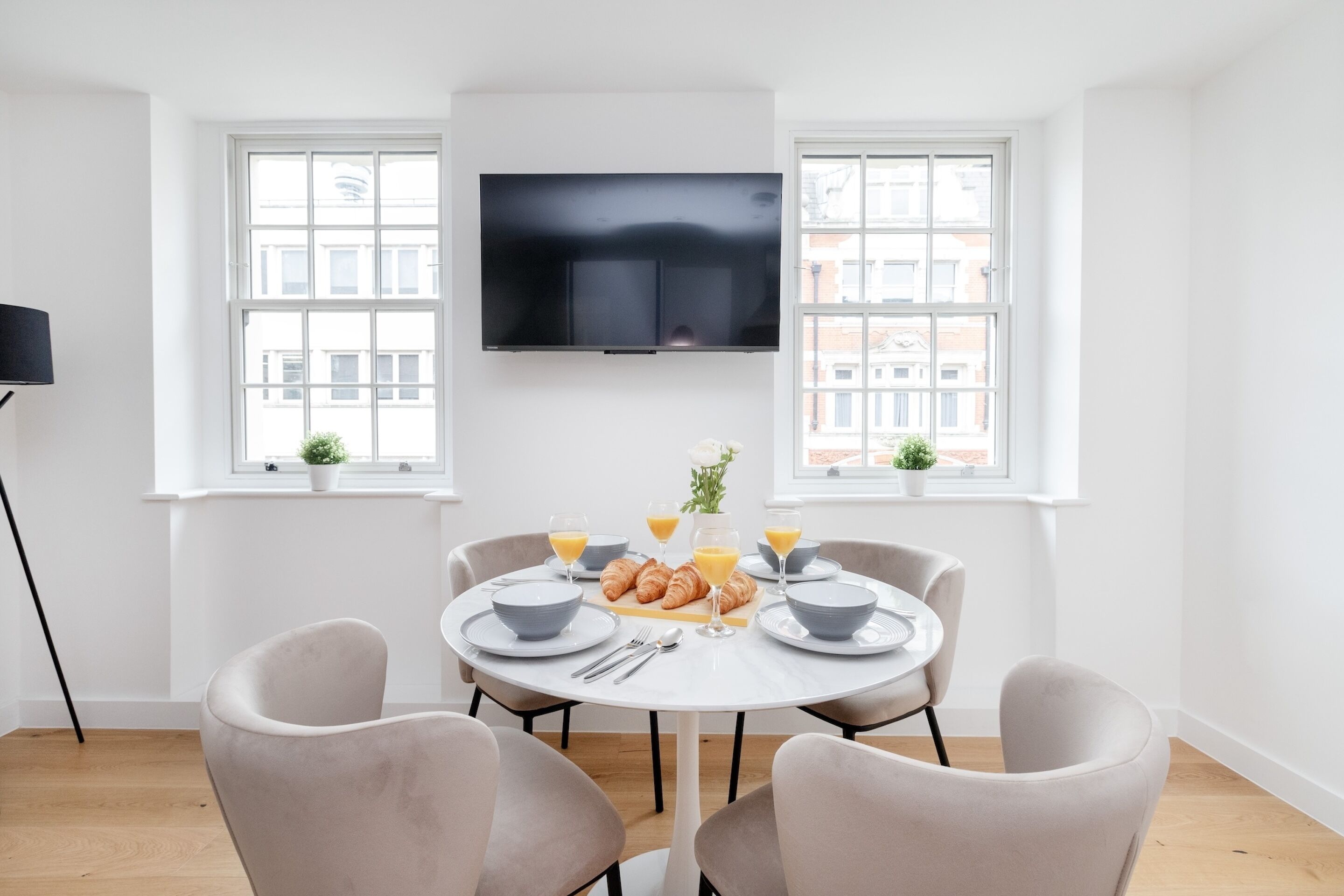 Een eethoek met een ronde tafel, vier stoelen, een flatscreen televisie aan de muur en twee ramen met potplanten.