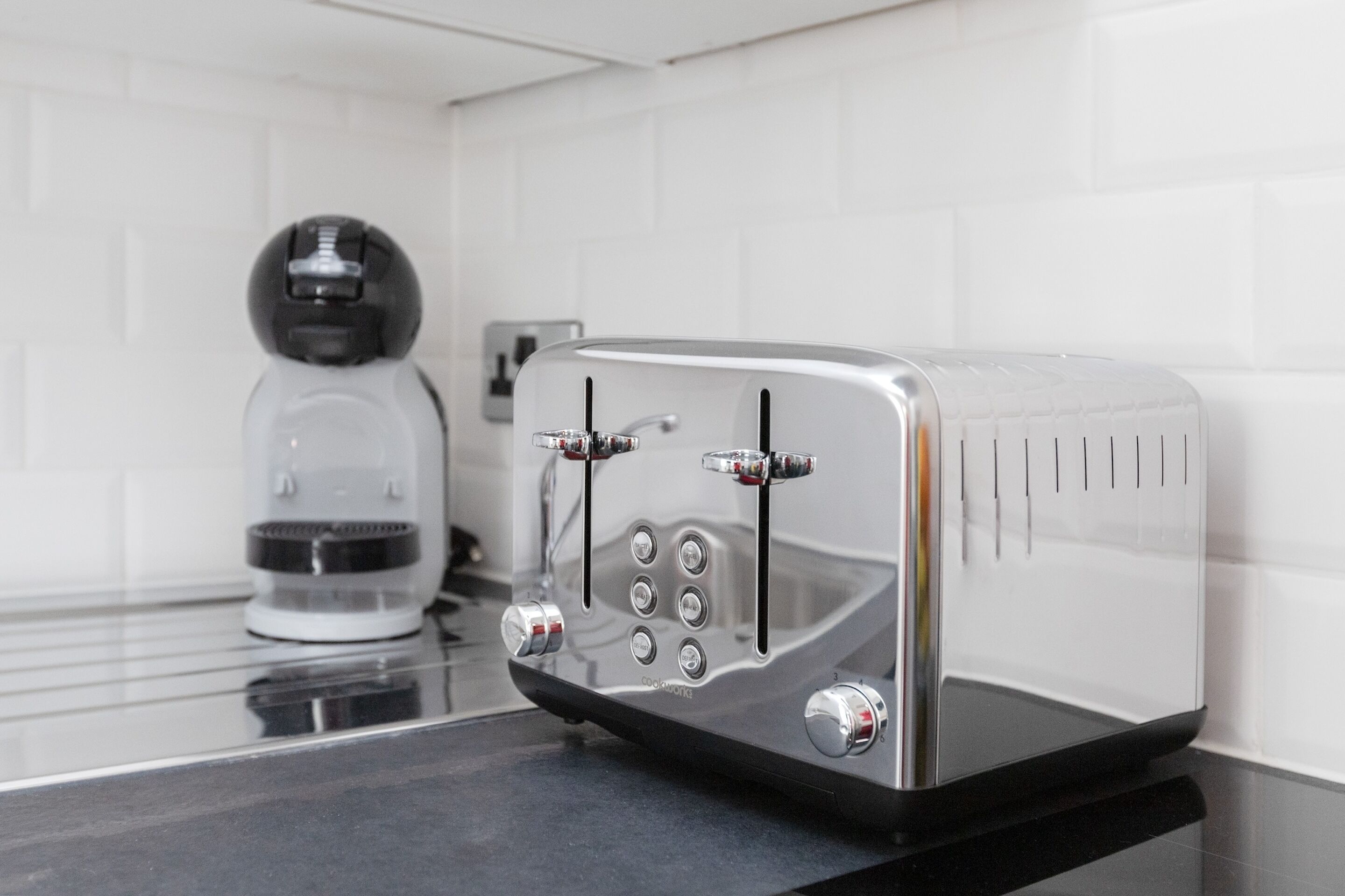 A modern kitchen with a stainless steel toaster on a black countertop, a coffee maker, and white tiled backsplash.