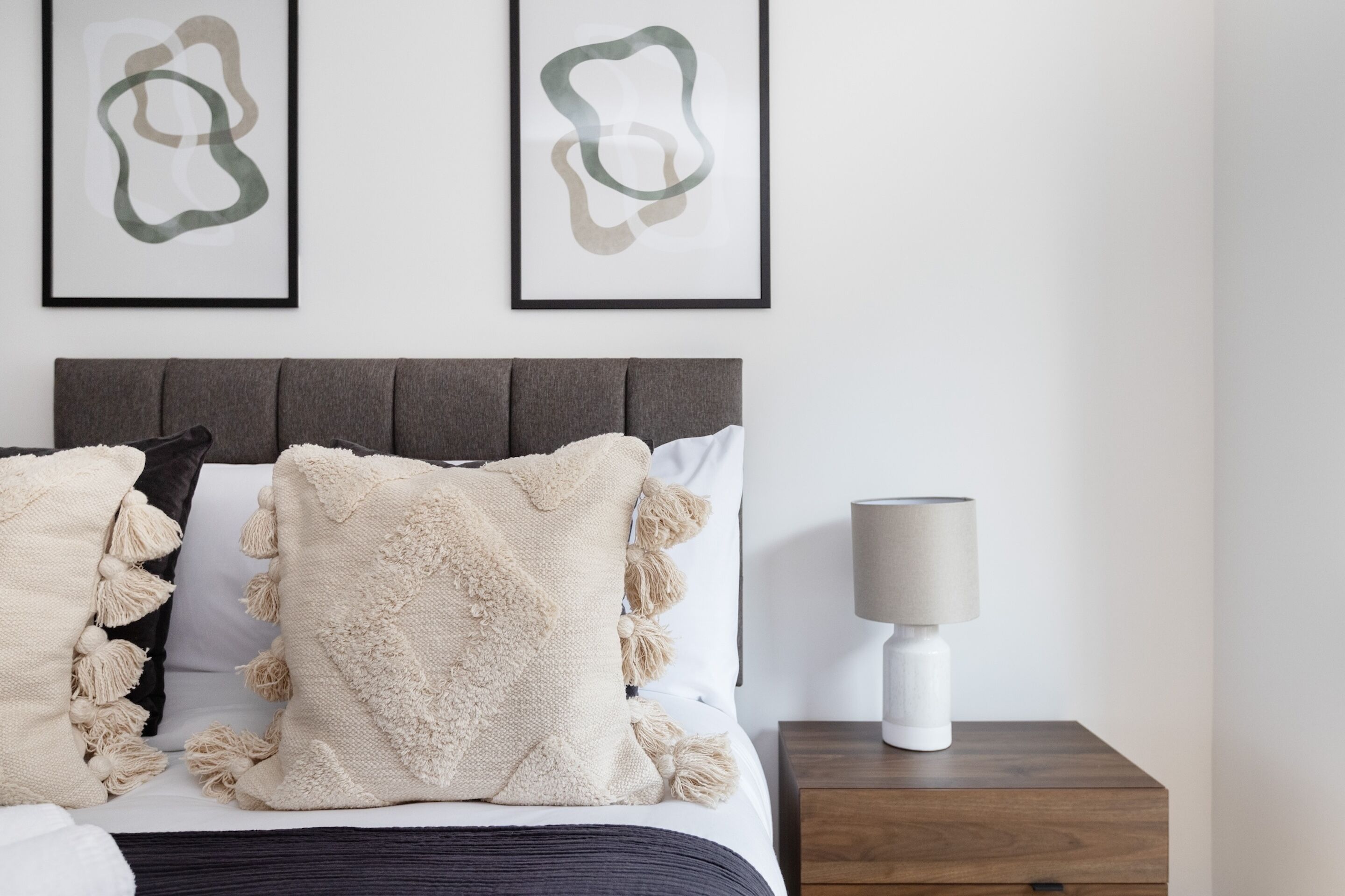 Ein ordentlich bezogenes Bett mit grauem Kopfteil, zwei gerahmte abstrakte Kunstwerke an der Wand und ein Nachttisch mit Lampe.