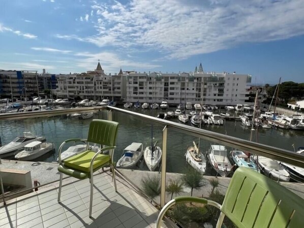 A marina with boats docked, a building with a pointed roof in the background, and a tiled deck with green chairs.