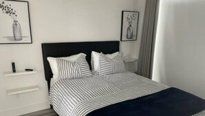 Ein ordentlich bezogenes Bett mit gestreifter Bettwäsche und einer dunkelblauen Tagesdecke, ein weißer Nachttisch mit einer kleinen Lampe sowie zwei gerahmte Bilder an der Wand.