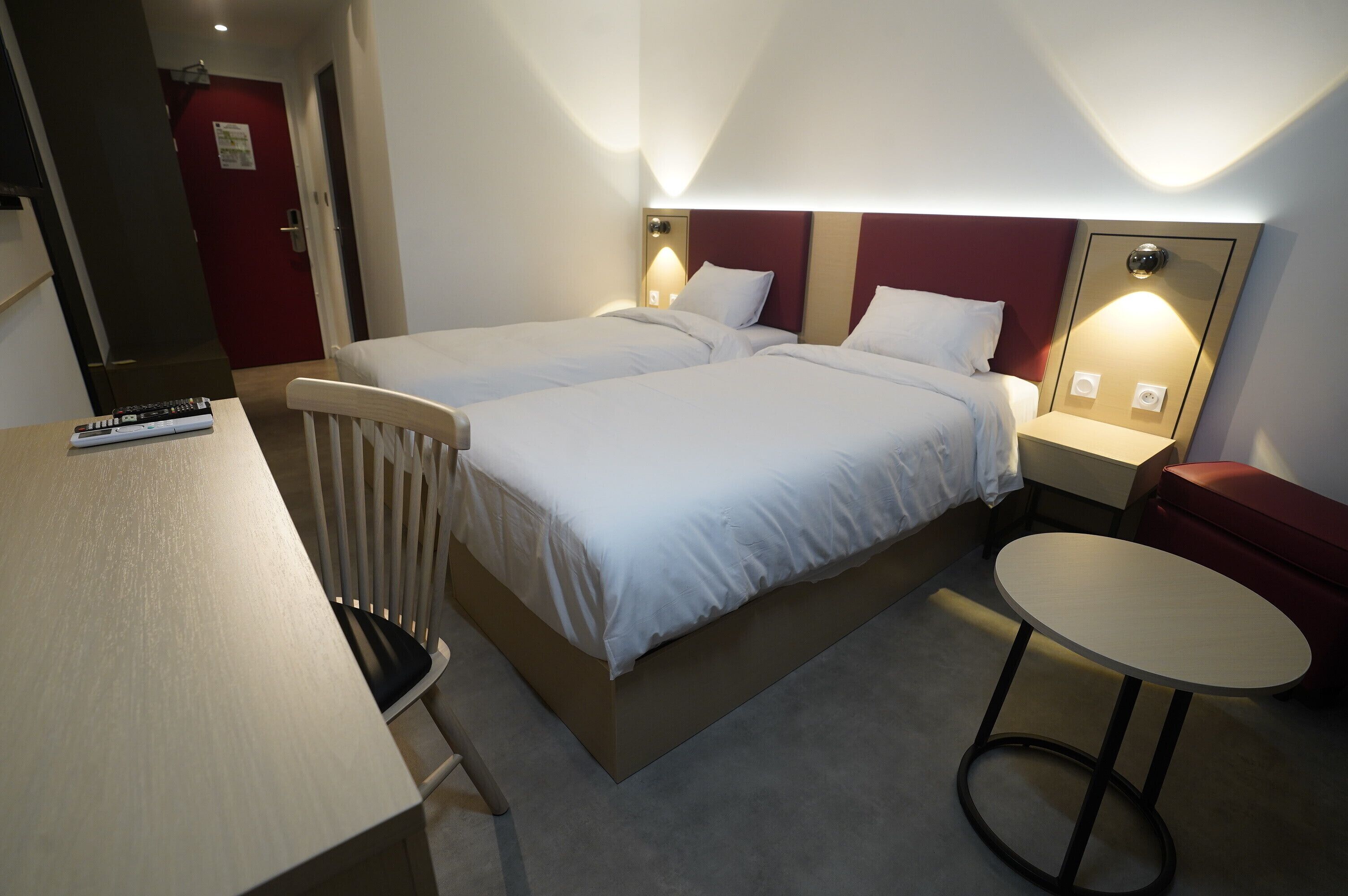 Habitación de hotel con dos camas, una mesita redonda pequeña, una silla y un escritorio con control remoto.