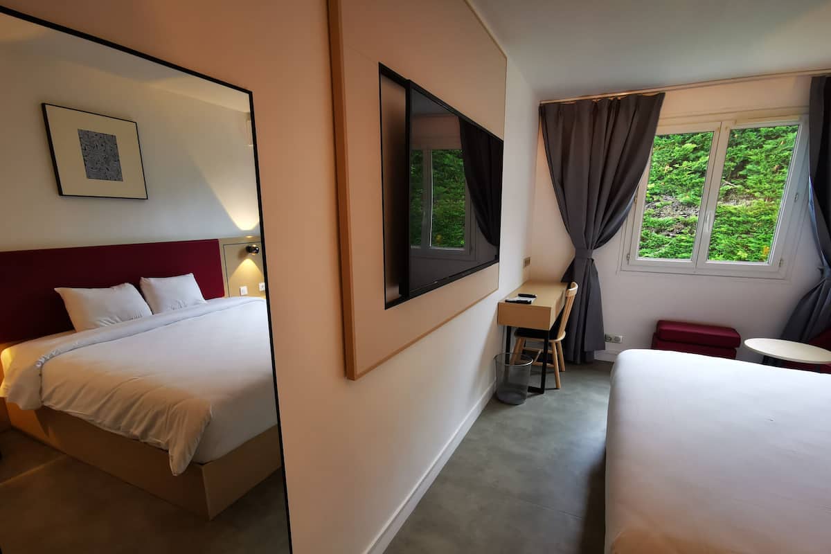 A hotel room with two beds, a desk, a chair, a small table, a trash bin, and a window with curtains.