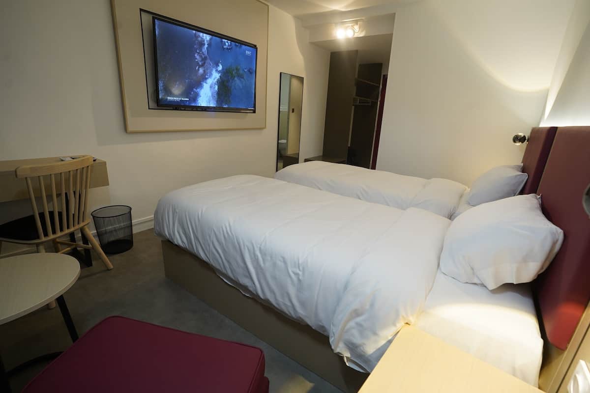 A hotel room with two beds, a TV mounted on the wall, a desk, and a chair.