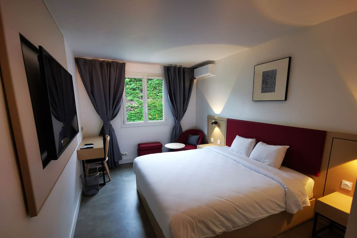 A hotel room with a bed, a desk, a chair, a small table, a window with curtains, and a wall-mounted air conditioner.