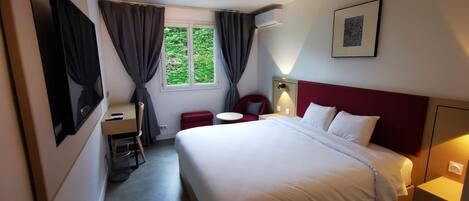 A hotel room with a bed, a desk, a chair, a small table, a window with curtains, and a wall-mounted air conditioner.