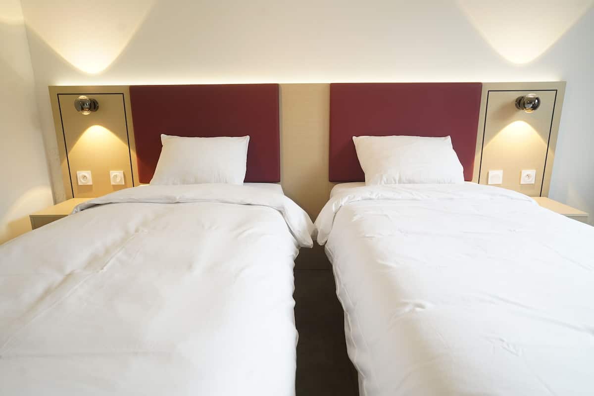 A hotel room with two beds, each with white linens and a red headboard. There are wall-mounted lamps on either side of the beds.