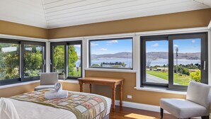 A bedroom with a large bed, two chairs, a small table, and multiple windows offering views of a lake and hills. - Coco's Place, your magical Snowy Mountains getaway. (Jindabyne)
