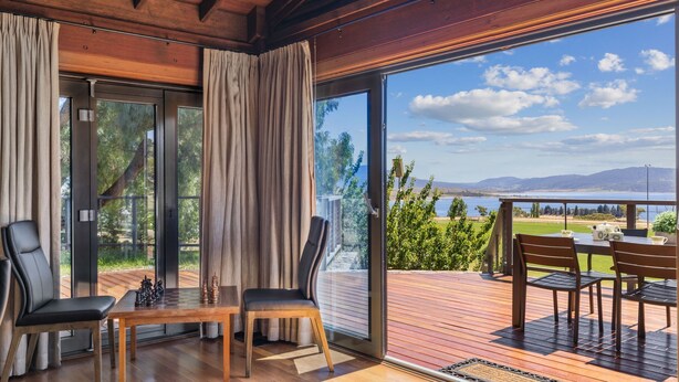 A room with a wooden deck, a table with chairs, and a view of a lake and mountains. - Coco's Place, your magical Snowy Mountains getaway. (Jindabyne)