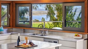 A kitchen with a large window overlooking a scenic landscape, a sink, and a countertop with a bottle and snacks. - Coco's Place, your magical Snowy Mountains getaway. (Jindabyne)