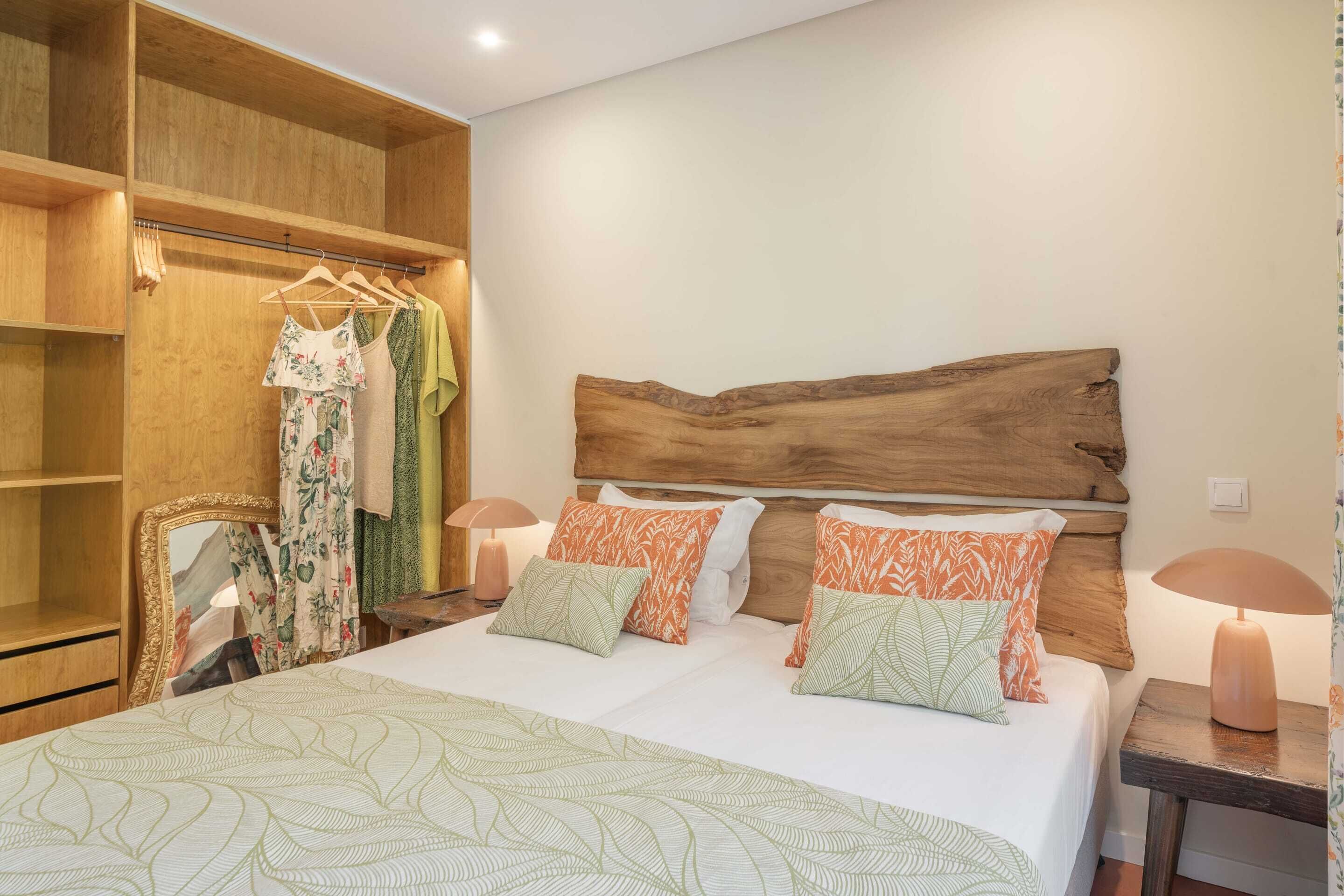 Une chambre à coucher avec une tête de lit en bois, un grand lit avec une literie à motifs, une armoire où sont suspendus des vêtements et des lampes de chevet.