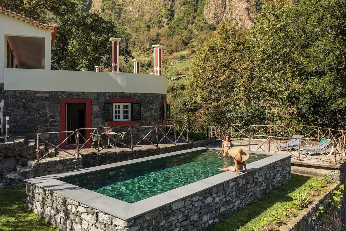 Een stenen zwembad met uitzicht op het omliggende landschap, een stenen gebouw met een rode deur, en een houten terras met lounge fauteuils.