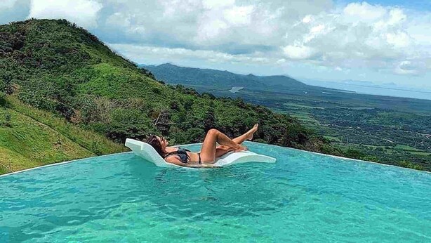 A person lying on a floating platform in a clear blue pool with a view of a green hillside and distant mountains.